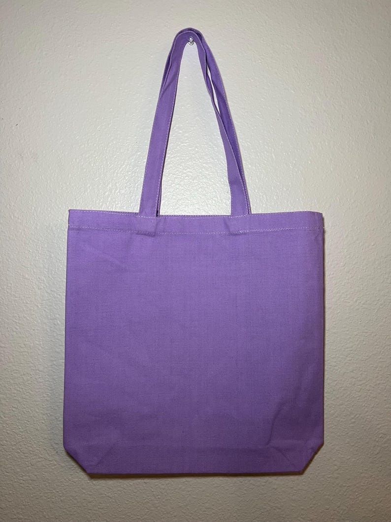 以下が含まれることがあります： 無地のラベンダー色のトートバッグで、お揃いのハンドルが付いています。丈夫な生地で作られており、物を運ぶように設計されています。ハンドルの耐久性を高めるために、バッグの上部に縫い付けられています。白い壁に掛けられています。