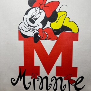 May include: White tote bag featuring a graphic of Minnie Mouse. Minnie Mouse is depicted in black, white, red, and yellow, resting on a large red letter "M". The name "Minnie" is written in black script below the letter.