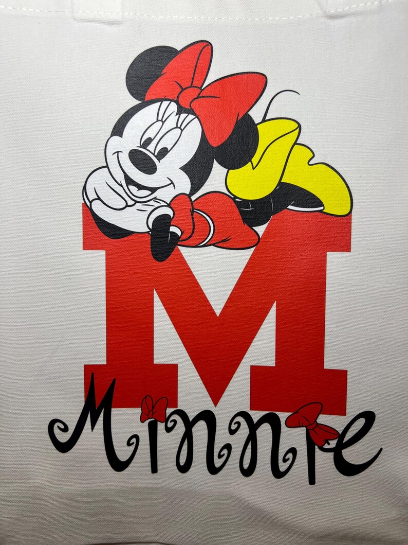 May include: White tote bag featuring a graphic of Minnie Mouse. Minnie Mouse is depicted in black, white, red, and yellow, resting on a large red letter "M". The name "Minnie" is written in black script below the letter.