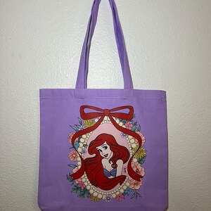 以下が含まれることがあります： ラベンダー色のトートバッグ。赤い髪の女性が紫色の貝殻のトップスを着て、花と真珠に囲まれたイラストがフレームに入っています。赤いリボンがフレームの上部を飾っています。バッグにはお揃いのラベンダー色のハンドルが付いています。