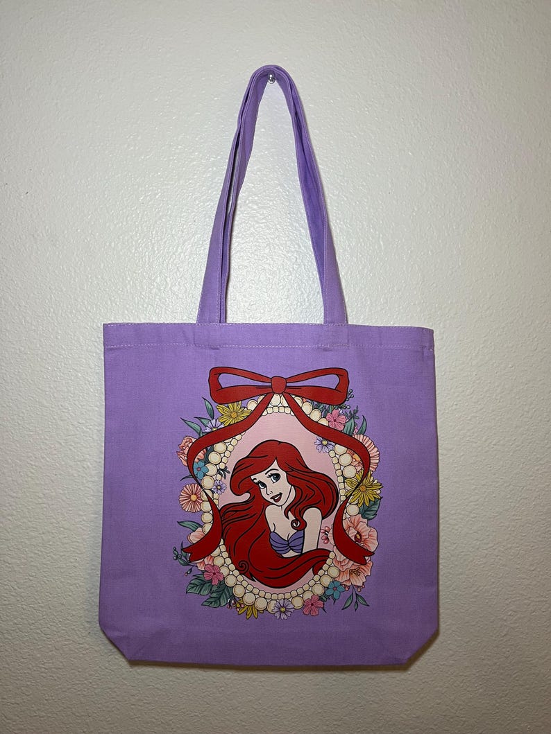 以下が含まれることがあります： ラベンダー色のトートバッグ。赤い髪の女性が紫色の貝殻のトップスを着て、花と真珠に囲まれたイラストがフレームに入っています。赤いリボンがフレームの上部を飾っています。バッグにはお揃いのラベンダー色のハンドルが付いています。