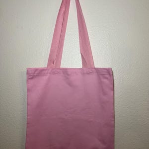 以下が含まれることがあります： 白い壁に、持ち手と一体型の無地のピンク色のトートバッグが吊り下げられています。バッグは長方形でシンプルなデザインで、様々なアイテムの持ち運びに適しています。持ち手は同じピンク色の生地で作られています。