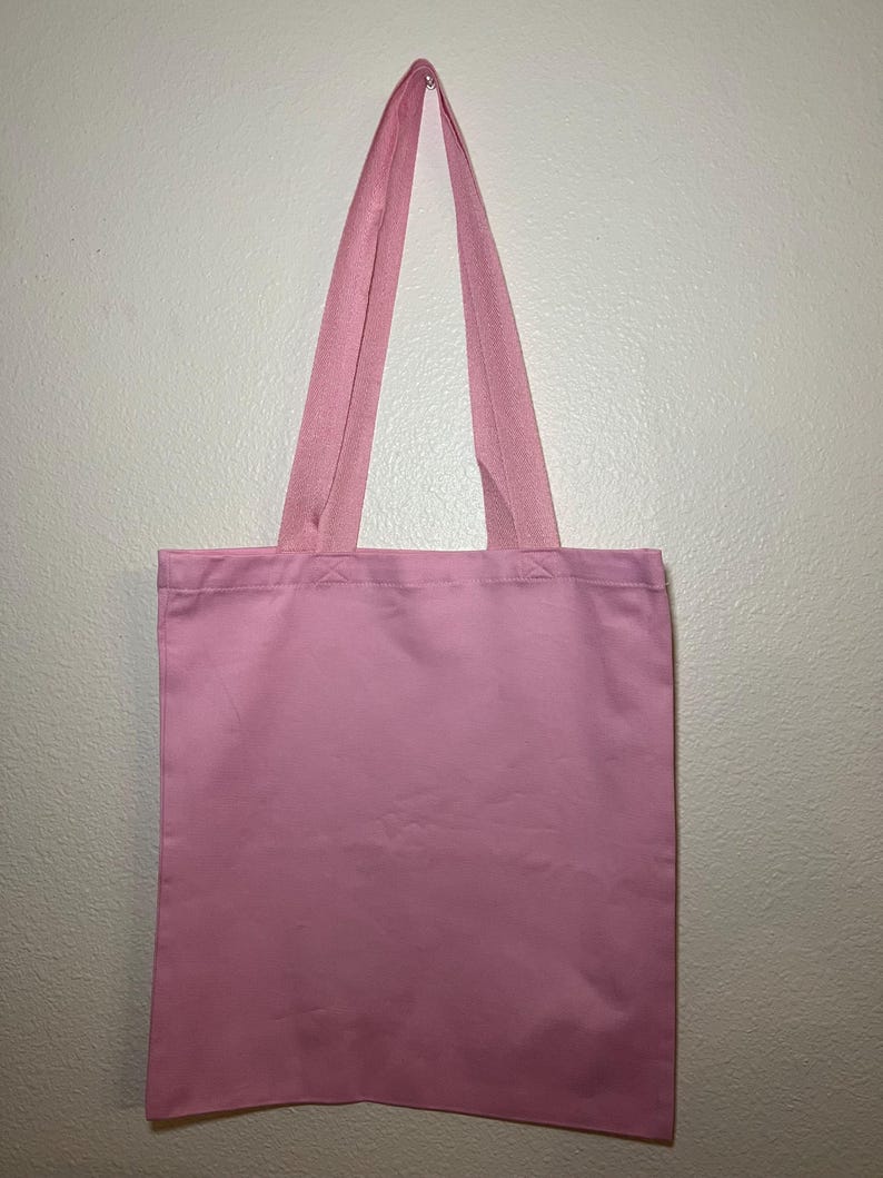 以下が含まれることがあります： 白い壁に、持ち手と一体型の無地のピンク色のトートバッグが吊り下げられています。バッグは長方形でシンプルなデザインで、様々なアイテムの持ち運びに適しています。持ち手は同じピンク色の生地で作られています。