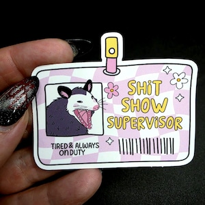 May include: A sticker with a pink and white checkered background, featuring a cartoon opossum with an open mouth. Text reads "Shit Show Supervisor" and "Tired & Always On Duty."