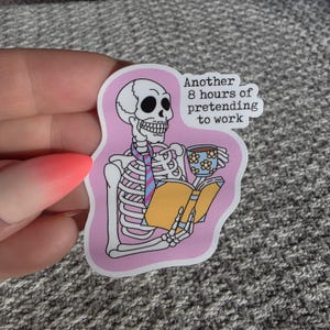 May include: A pink sticker with a cartoon skeleton reading a book and drinking from a floral-patterned mug. The text reads "Another 8 hours of pretending to work."