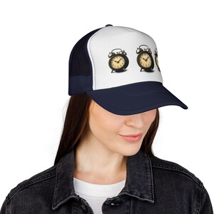 May include: A navy blue and white trucker hat featuring a design of three vintage alarm clocks. The clocks are black with gold-colored faces and hands. The hat has a curved brim and a mesh back.
