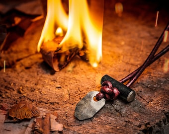 May include: A close-up of a fire starter with a dark brown leather cord and a metal striker. The fire starter is sitting on a wooden surface with a fire burning in the background.
