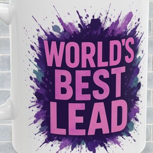 以下が含まれることがあります： 「WORLD'S BEST LEAD」のピンク色の文字が特徴の白いセラミックマグ。紫とターコイズのスプラッシュデザインで囲まれています。マグには湾曲したハンドルが付いています。背景は灰色のレンガの壁です。