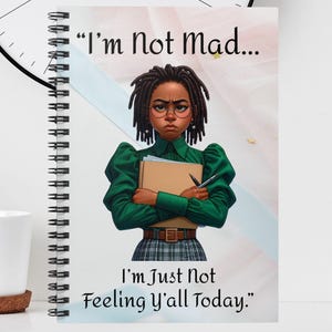 May include: A spiral-bound notebook with the text "I'm Not Mad... I'm Just Not Feeling Y'all Today." features an illustration of a woman in a green top and plaid skirt. A white mug, vase with flowers, and a black and white clock are also in the image.