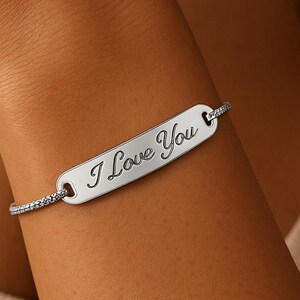 May include: A silver bracelet with the inscription "I Love You" on a rectangular plate. The bracelet features a delicate chain with small, sparkling accents. The bracelet is worn on a wrist.