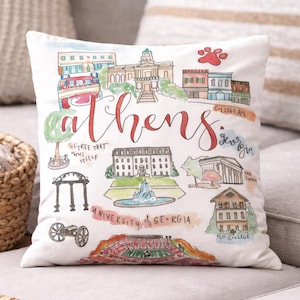 May include: White decorative pillow featuring a watercolor illustration of Athens, Georgia landmarks. The design includes the word "Athens" in red script, along with illustrations of buildings, a paw print, and the text "College Ave" and "University of Georgia."