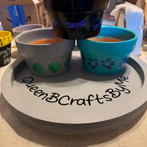 May include: Three painted flower pots in black, gray, and teal, stacked on a gray tray. The black pot has a purple dragonfly design, the gray pot has green leaf designs, and the teal pot has purple flower designs. The tray has the text "QueenBCraftsByMe".