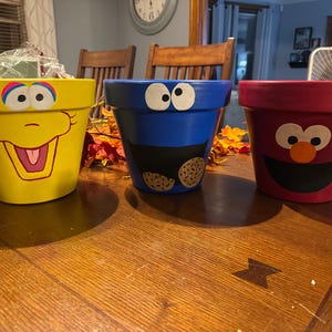 May include: Three hand-painted flower pots featuring Sesame Street characters. The yellow pot depicts a smiling face with a large open mouth. The blue pot has a cookie monster design, and the red pot has an Elmo design.