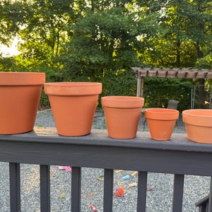 May include: A set of five terracotta plant pots arranged in a row by size, from largest to smallest. Each pot features a classic tapered shape with a wide rim at the top and a smooth, unglazed orange-brown surface, showcasing a consistent design.
