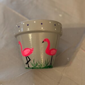 May include: A gray painted terracotta flower pot with a pink flamingo design. The pot features two flamingos, green grass, and white and purple polka dots around the rim. The flamingos are painted in bright pink with black legs and beaks.
