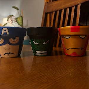 May include: Three hand-painted terracotta flower pots featuring superhero designs. The pots are painted with Captain America, Hulk, and Iron Man faces. The Captain America pot is blue with a white "A", the Hulk pot is green, and the Iron Man pot is gold and red.