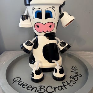 May include: A decorative cow figure crafted from stacked terracotta pots painted with black and white spots. The figure features a painted face with blue eyes and a pink snout, with smaller pots as limbs. The text QueenBCraftsByMe is visible.