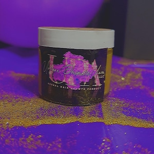 May include: A jar of herbal hair growth product with a white lid and a black label. The label features the text "Unique Primrose Ash LLC" in a stylized font, with a pink floral design. The jar sits on a purple surface with gold glitter.