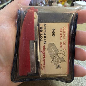 May include: A vintage stapler and staple set in a clear plastic pouch. The red stapler has silver details, and the cream-coloured staple box features red text reading "Swingline" and "1000 Staples Chisel Pointed".