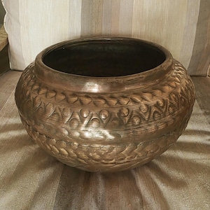 May include: A round, metallic bowl with a dark bronze finish. The exterior features a wide, horizontal band of embossed circular and crescent-shaped patterns, complemented by textured, scale-like detailing around the lower section.
