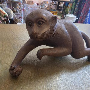 May include: A brown wooden monkey sculpture in a crawling pose. The monkey has detailed facial features and is holding a small ball in its hand. The sculpture is on a textured surface.