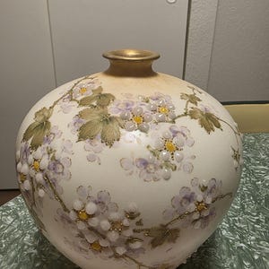 Könnte beinhalten: Eine runde, cremefarbene Keramikvase mit einem erhabenen, handbemalten Blumenmuster aus weißen und hellvioletten Blütenblättern, gelben Zentren und grünen Blättern. Die Vase hat einen goldfarbenen Rand und einen bauchigen Körper.