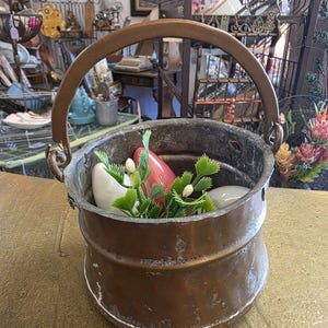 Puede incluir: Un pequeño cubo de cobre antiguo con un asa curvada. El cubo contiene piedras decorativas blancas y rojas y ramitas verdes. El cobre tiene una pátina desgastada, lo que sugiere edad y uso. El cubo es probablemente para fines decorativos.