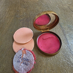 May include: Vintage compact with a gold-toned, ribbed case, holding pink blush and a mirror. Includes three peach-colored powder puffs and a small, round, silver-colored item with the text "Evening in Paris".
