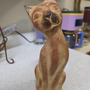 May include: A tan-colored ceramic cat figurine with a slightly tilted head and a smiling expression. The cat has detailed facial features and a slender body. The figurine is a decorative item.