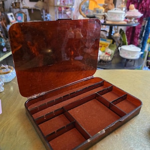 May include: Open, vintage jewelry box with a tortoiseshell finish. The box is rectangular with a hinged lid and a series of compartments lined with a reddish-brown material. The interior is divided into various sections for organizing jewelry.