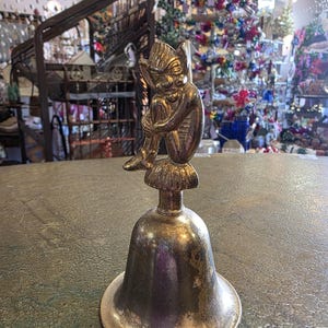 Könnte beinhalten: Eine silberfarbene Vintage-Glocke mit einer sitzenden Elfenfigur obenauf. Der Elf ist goldfarben und scheint in Gedanken versunken zu sein. Die Glocke hat einen ausgestellten Sockel und eine angelaufene Oberfläche, die auf das Alter hindeutet. Der Hintergrund ist ein Geschäftsumfeld.