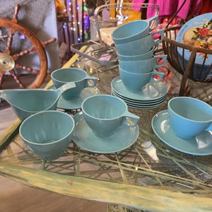 May include: A collection of vintage aqua-coloured teacups and saucers. The set includes several cups, saucers, and a creamer, all in a matching light blue hue. The cups have a unique handle design. A decorative floral tin is in the background.