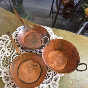 May include: Three antique copper pans and a small copper cup with handles rest on a white lace doily. The pans have long wooden handles, and the cup has a small metal handle. The copper has a warm, aged patina.