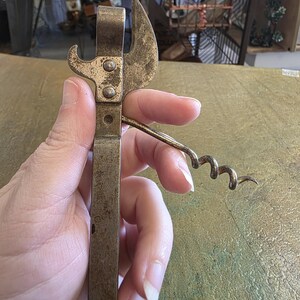 May include: Vintage metal combination can opener and corkscrew. The opener is a tarnished brass color, with a hook for opening cans and a corkscrew for removing corks from bottles. The tool is held in a hand.