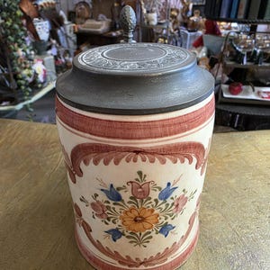 Könnte beinhalten: Ein zylindrischer Bierkrug aus Keramik mit einem dunkelgrauen Metalldeckel und einem dekorativen Knauf. Der cremefarbene Körper ist mit handgemalten Blumenmotiven in Orange, Blau und Rosa verziert, die von rotbraunen Zierrändern umrahmt sind.