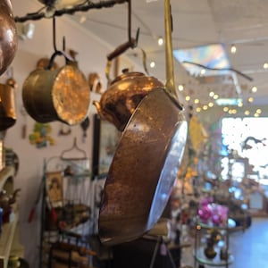 Könnte beinhalten: Eine kupferfarbene Bratpfanne mit langem Messinggriff hängt an einem Metallhaken. Weitere Kupferkochutensilien, darunter Töpfe und ein Wasserkocher, sind im Hintergrund aufgehängt. Das Kochgeschirr hat eine Vintage-Ästhetik.