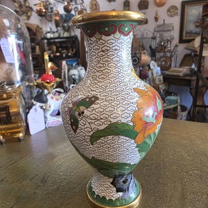 Pode incluir: Um vaso decorativo com aro e base dourados. O vaso apresenta um fundo branco com um padrão de nuvens repetido. Possui um design floral com detalhes em laranja, verde e vermelho. Uma borboleta também é visível no vaso.