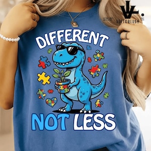 Könnte beinhalten: Blaues T-Shirt mit der Aufschrift "DIFFERENT NOT LESS" und einem Cartoon-Dinosaurier mit Sonnenbrille. Der Dinosaurier hält eine Pflanze in einem Topf, der mit Puzzleteilen verziert ist. Bunte Puzzleteile sind verstreut.