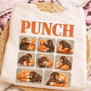 May include: White sweatshirt with the word "PUNCH" in orange block letters. Below the text are nine squares, each with a picture of two hugging monkeys. The monkeys are brown and orange.