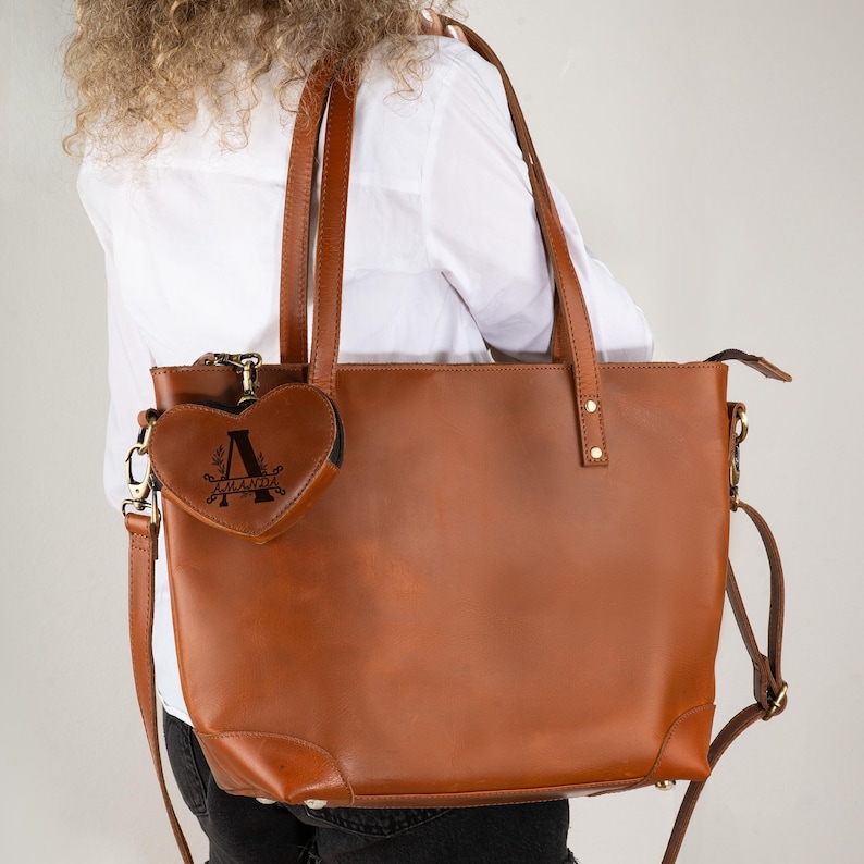 May include: A brown leather tote bag with a matching heart-shaped coin purse. The tote has two top handles and a detachable shoulder strap. The coin purse is attached to the tote with a small chain. The coin purse has the name "AMANDA" and the letter "A" on it.