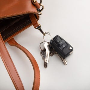 May include: A close-up of a brown leather handbag with a zipper and a strap. Attached to the bag is a keyring with several keys and a black car key fob. The bag and keys are on a white background.