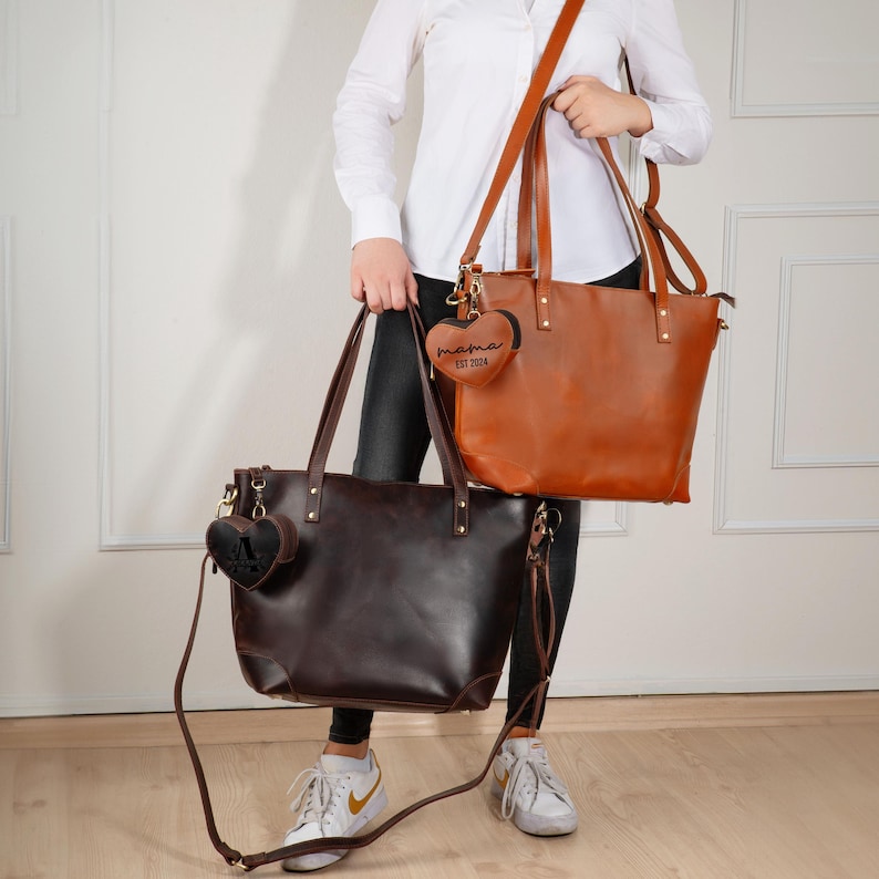 May include: Two leather tote bags in brown and dark brown, each with a long shoulder strap and a smaller heart-shaped charm. The charms read "mama EST 2024" and have a letter "A" on them. The bags have gold-colored hardware.