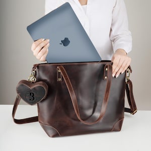 May include: A brown leather tote bag with a matching shoulder strap and a heart-shaped charm. A gray laptop is being placed inside the bag. The charm has a floral design and the letter "B".
