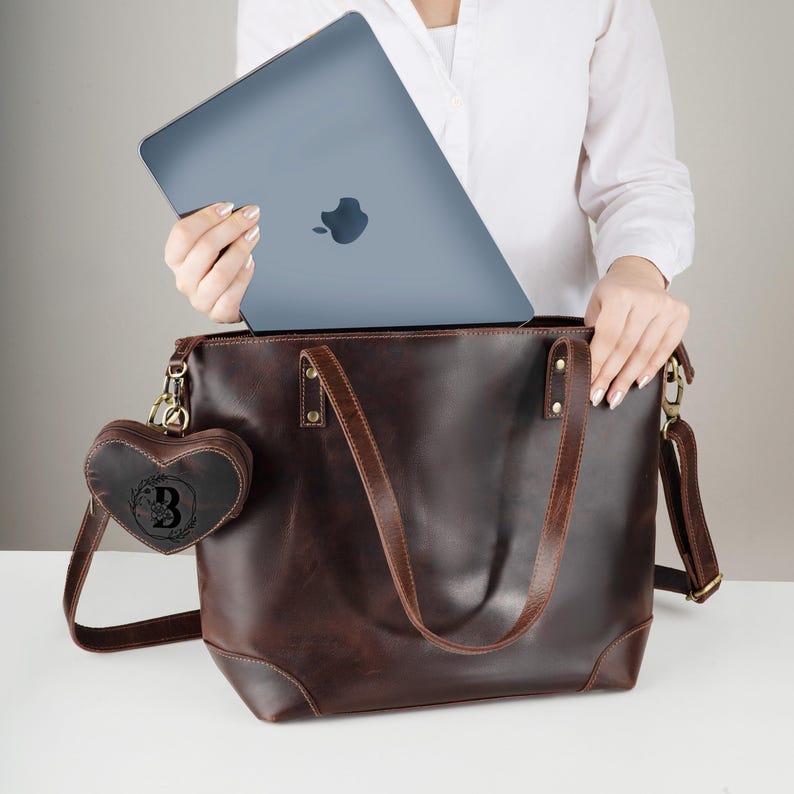 May include: A brown leather tote bag with a matching shoulder strap and a heart-shaped charm. A gray laptop is being placed inside the bag. The charm has a floral design and the letter "B".