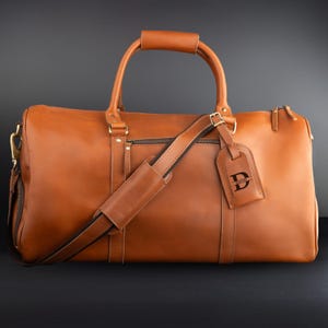 May include: A brown leather duffel bag with a top handle, shoulder strap, and a luggage tag. The bag has a zipper closure and gold-colored hardware. The luggage tag has the letter "D" on it.