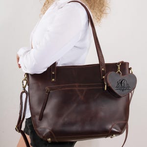 May include: A dark brown leather tote bag with a matching heart-shaped coin purse attached. The tote has a shoulder strap and a front zipper pocket. The coin purse has the name "Carolina" engraved on it.