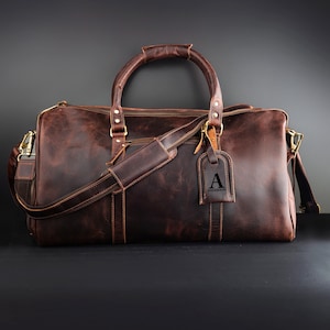 May include: A brown leather duffel bag with a top handle, shoulder strap, and a luggage tag. The bag has a zipper closure and gold-colored hardware. The luggage tag has the letter "A" and the name "ANDERSON" printed on it.