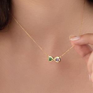 May include: A gold-colored necklace featuring an infinity symbol pendant. The pendant is adorned with a green and a purple gemstone. The necklace is delicate and appears to be worn around the neck.