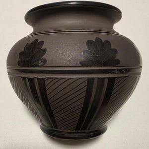 May include: A matte charcoal gray ceramic vase with a rounded body and a flared rim. The upper section features black painted floral motifs, while the lower section displays alternating black vertical bands and fine diagonal etched line patterns.