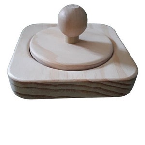 May include: A wooden toy with a round knob on top and a square base. The toy is made of light-colored wood and has a smooth finish.